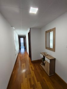 Pasillo largo con suelo de madera, paredes blancas, mueble recibidor con espejo y luz de techo en piso de alquiler en Carball - Serdava