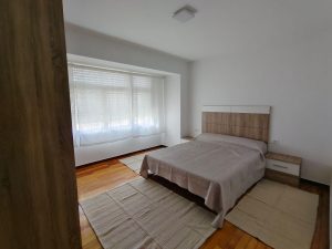 Dormitorio luminoso con cama de matrimonio, suelo de madera y grandes ventanales en piso de alquiler en Carballo - Serdava
