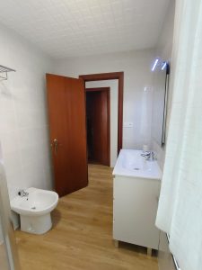 Cuarto de baño con suelo de madera, mueble de lavabo blanco, bidé y puertas de madera en piso de alquiler en Carballo - Serdava