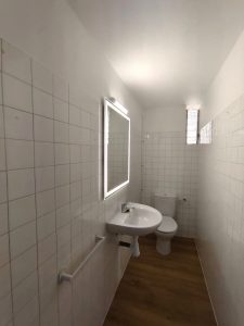Baño de piso en alquiler en Carballo con paredes de azulejos blancos, lavabo con espejo retroiluminado y suelo de madera - Serdava