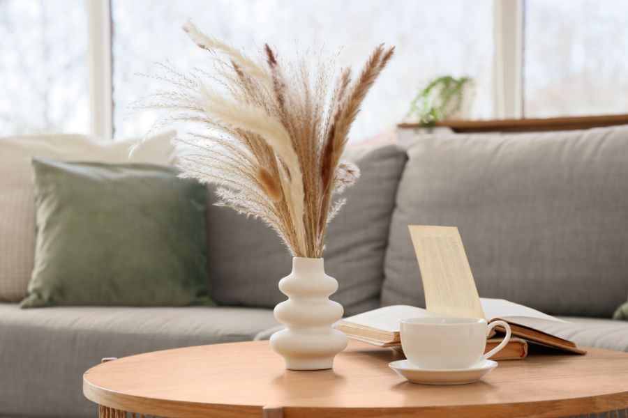 Jarrón con hierba de la pampa, libro abierto y taza de café sobre mesa de madera frente a un sofá gris - Serdava